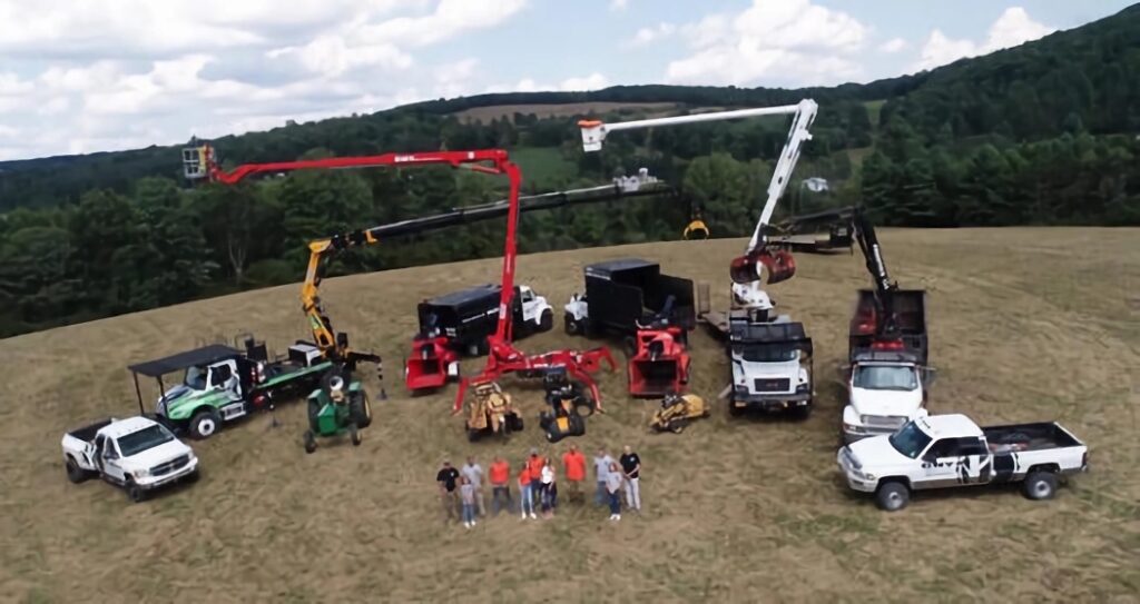 An aerial view showcasing the extensive fleet of tree service equipment owned by CNY Tree Service. The image features various specialized machinery, including bucket trucks, cherry pickers, stump grinders, and wood chippers, lined up in a field with a scenic forested backdrop. The team of six employees stands proudly in front of the equipment, wearing matching orange shirts, emphasizing professionalism and expertise.